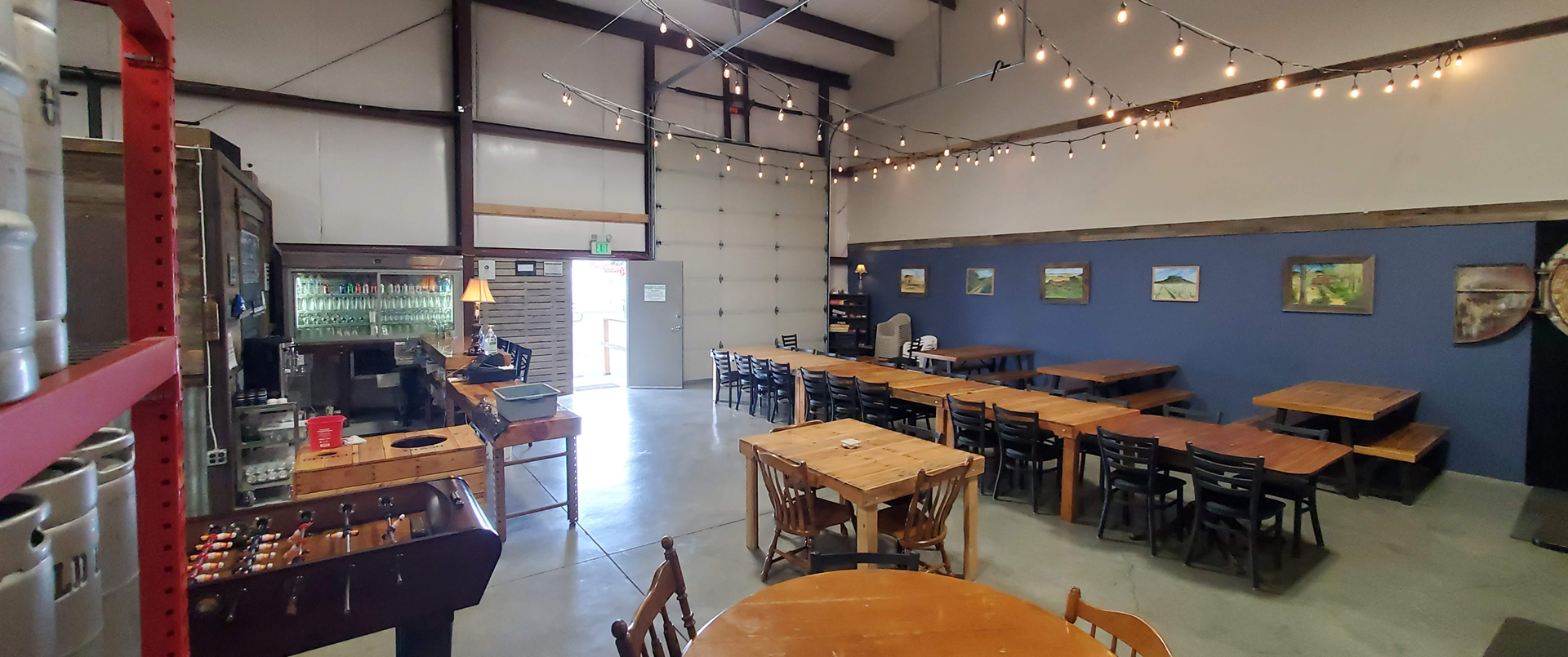 The tasting room is located in an industrial metal building wtih concrete floors. It is decorated with blue walls with landscape paintings. Cafe lights are strung over wooden tables.